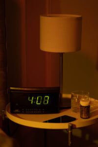 A warm and moody bedside table with a digital clock showing 4:08 and a lamp in dim lighting.